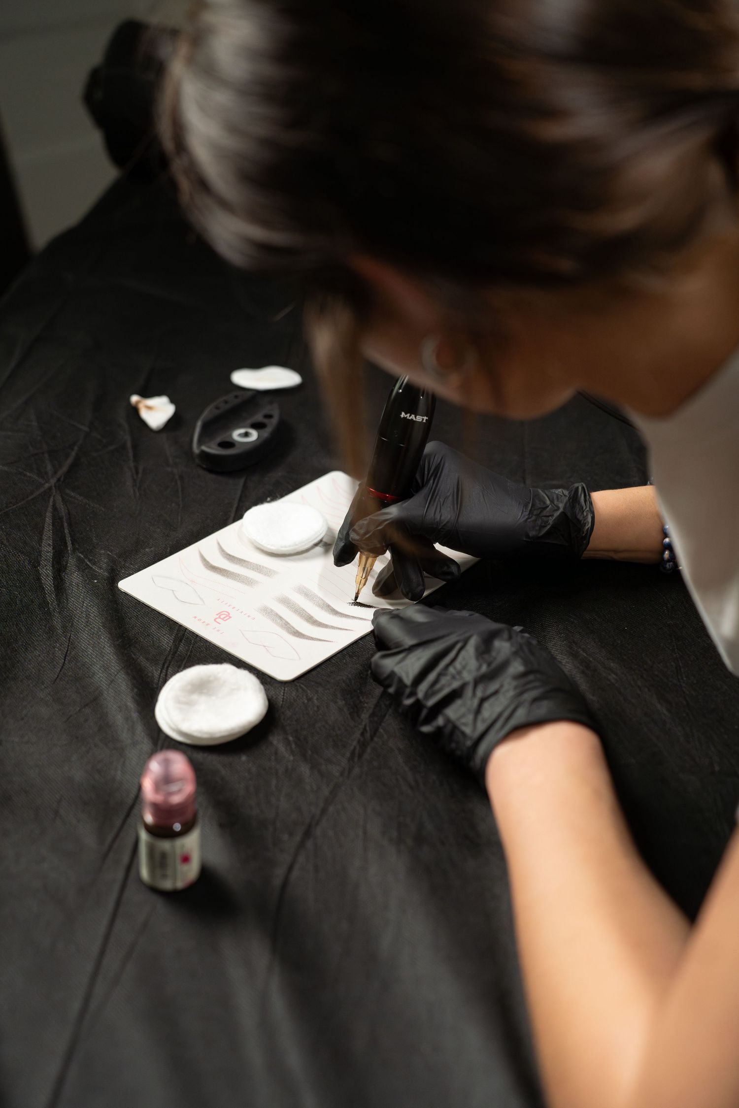 Beauty technician in black gloves practicing permanent makeup techniques on practice pad.