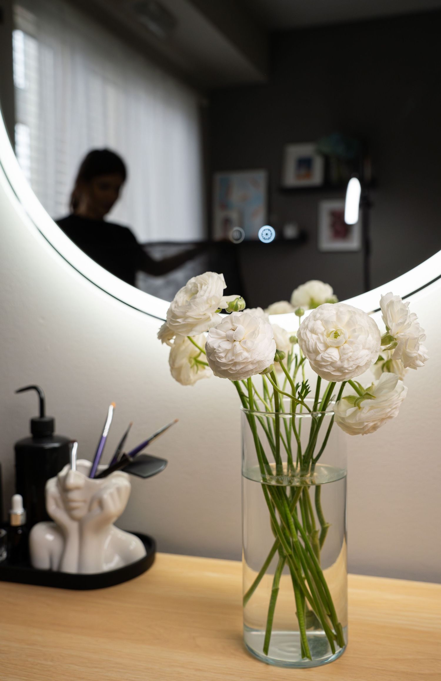 White flowers in glass vase reflected in round mirror with beauty supplies visible on counter.