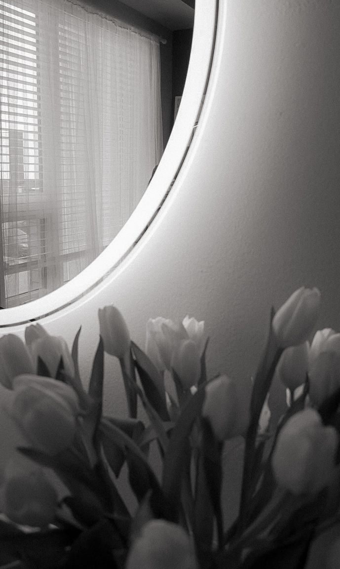 Black and white image of tulips by a curved window with blinds casting shadows.