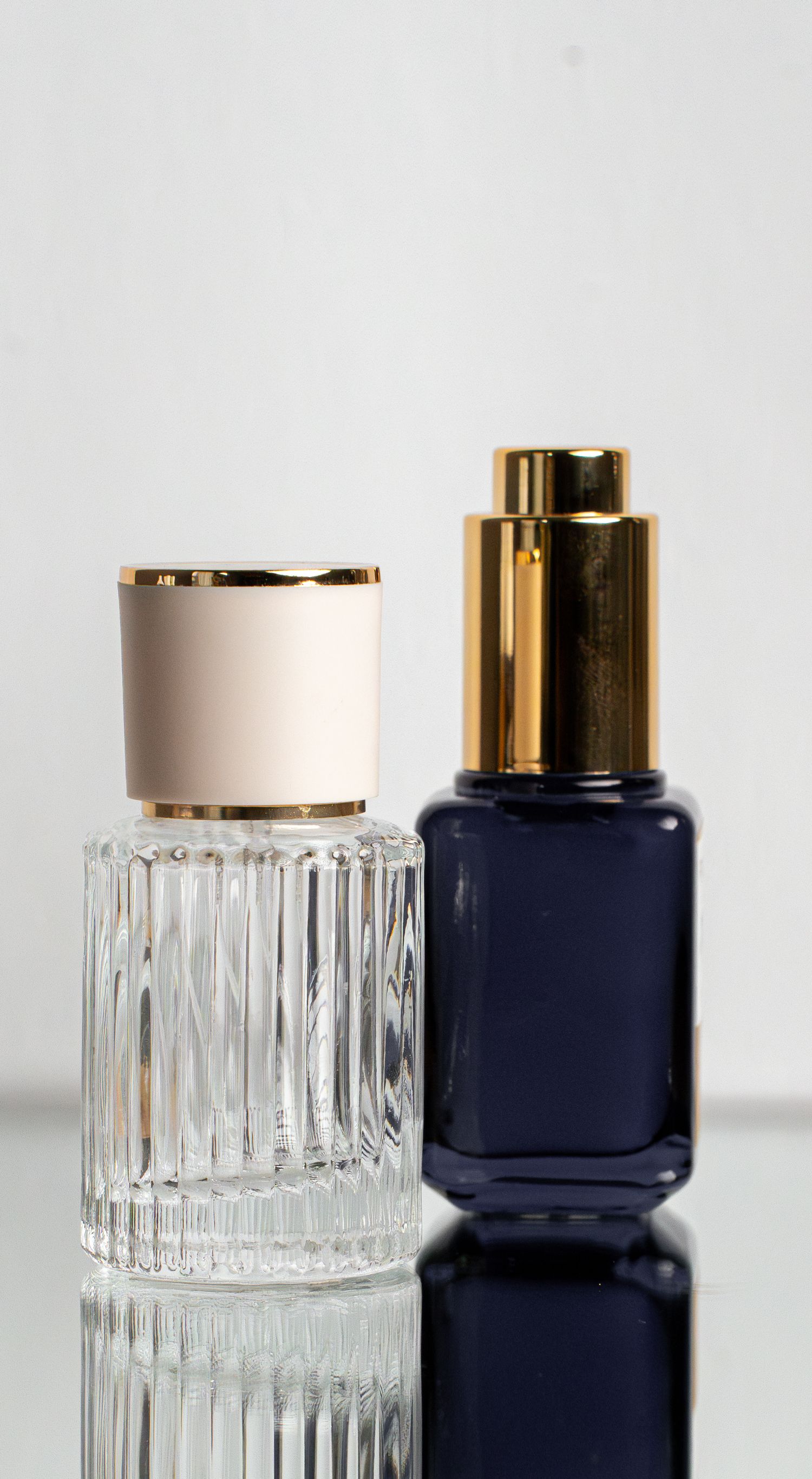 Two cosmetic bottles with gold caps, one clear glass with a ribbed texture and one navy blue, displayed on a reflective surface.