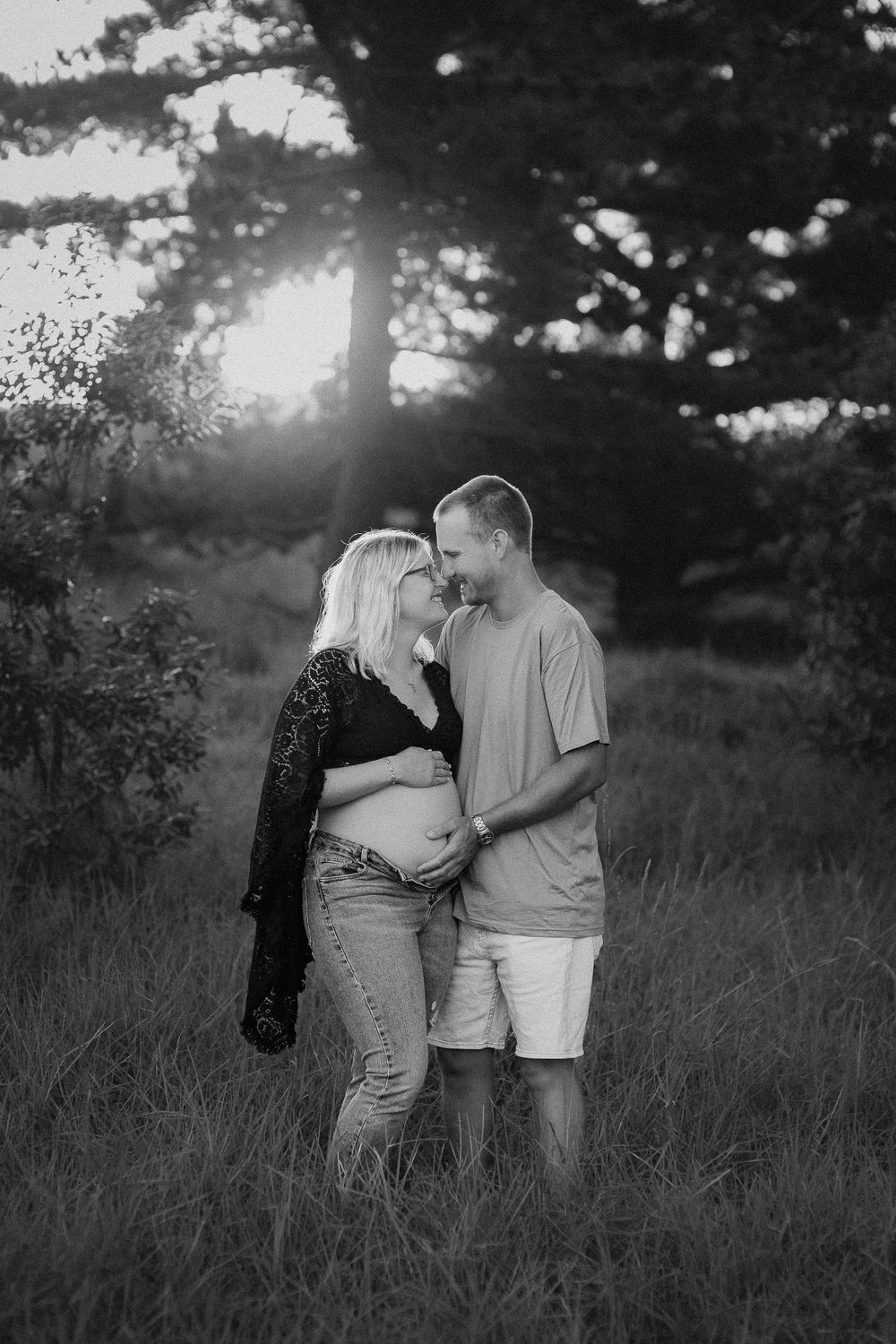 A couple embraces in a field during sunset with sunlight filtering through trees in black and white photography. Whakatane Photographer