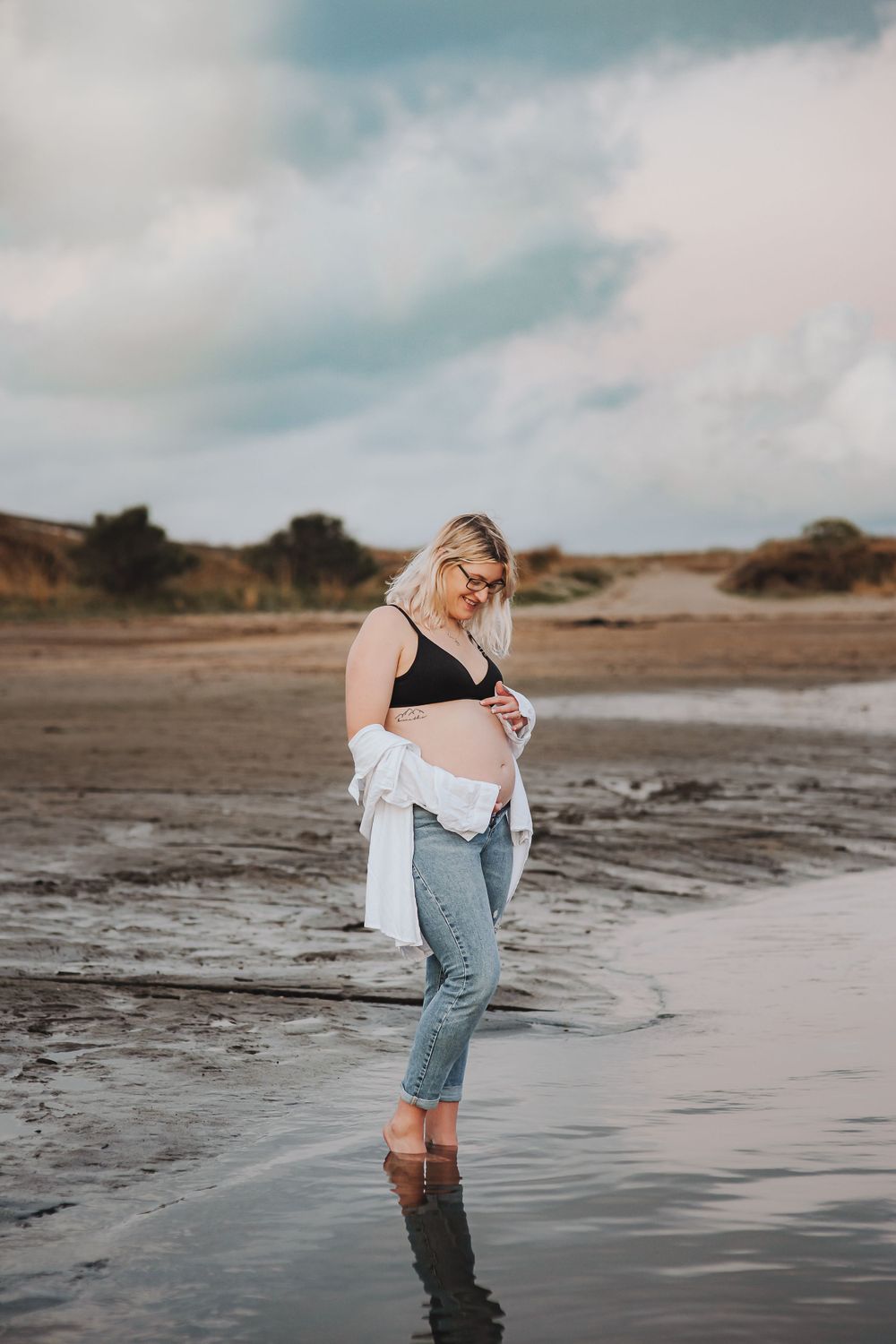 Beach maternity portrait with cloudy sky and woman standing in shallow water.