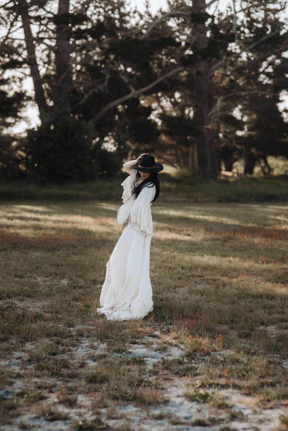 A figure in a flowing white vintage dress and dark wide-brimmed hat stands in a sunlit meadow surrounded by tall trees. Whakatane Maternity