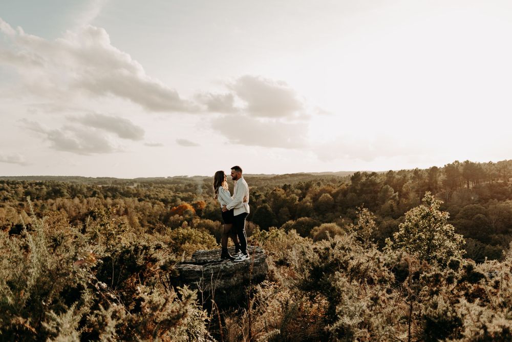 Two people stand together on a rocky cliff overlooking an autumn forest landscape at sunset.