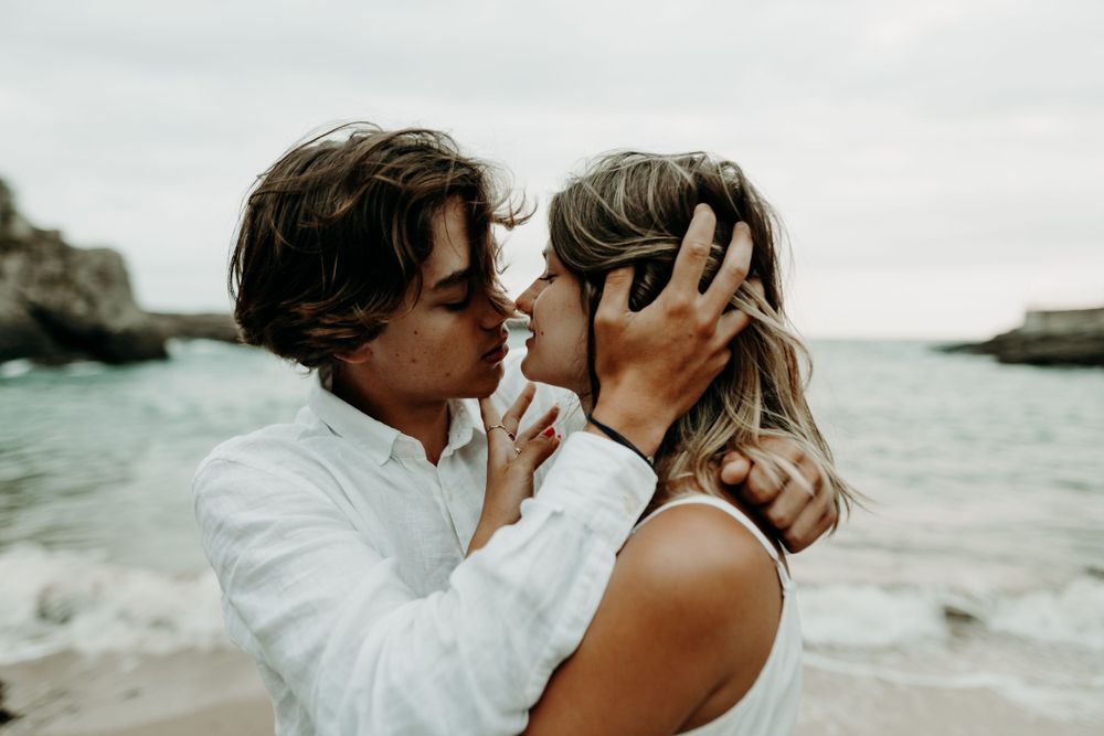 A romantic moment between a couple embracing on a rocky beach with ocean waves and cliffs in the background.