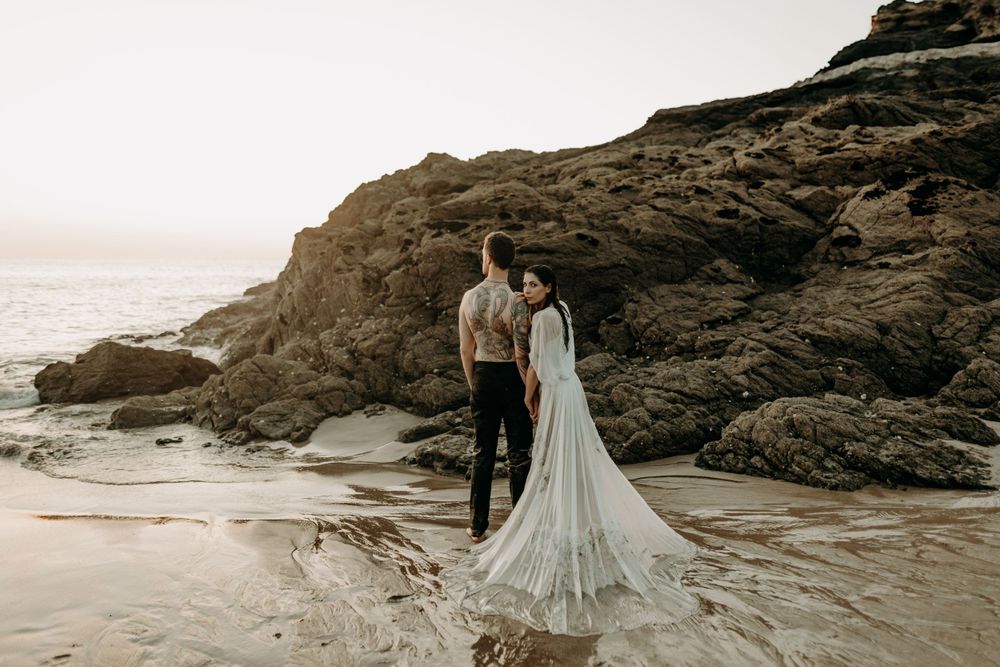 A couple in wedding attire stands on a rocky beach at sunset with waves crashing behind them.