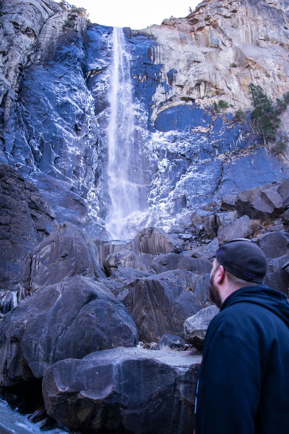Towering waterfall cascades down rocky cliff face with large boulders in foreground.