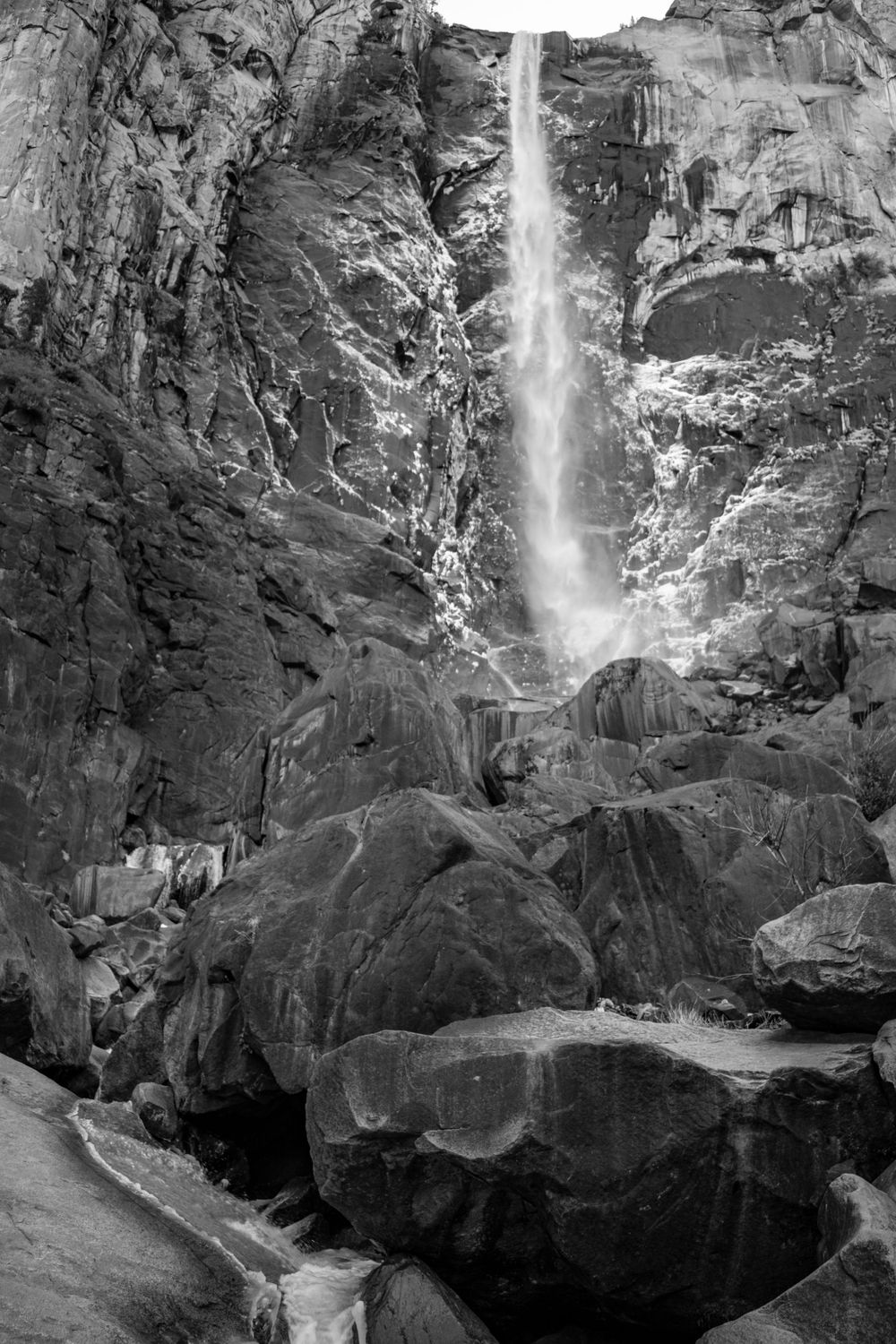 Black and white capture of waterfall streaming down steep rocky cliff with boulders below.