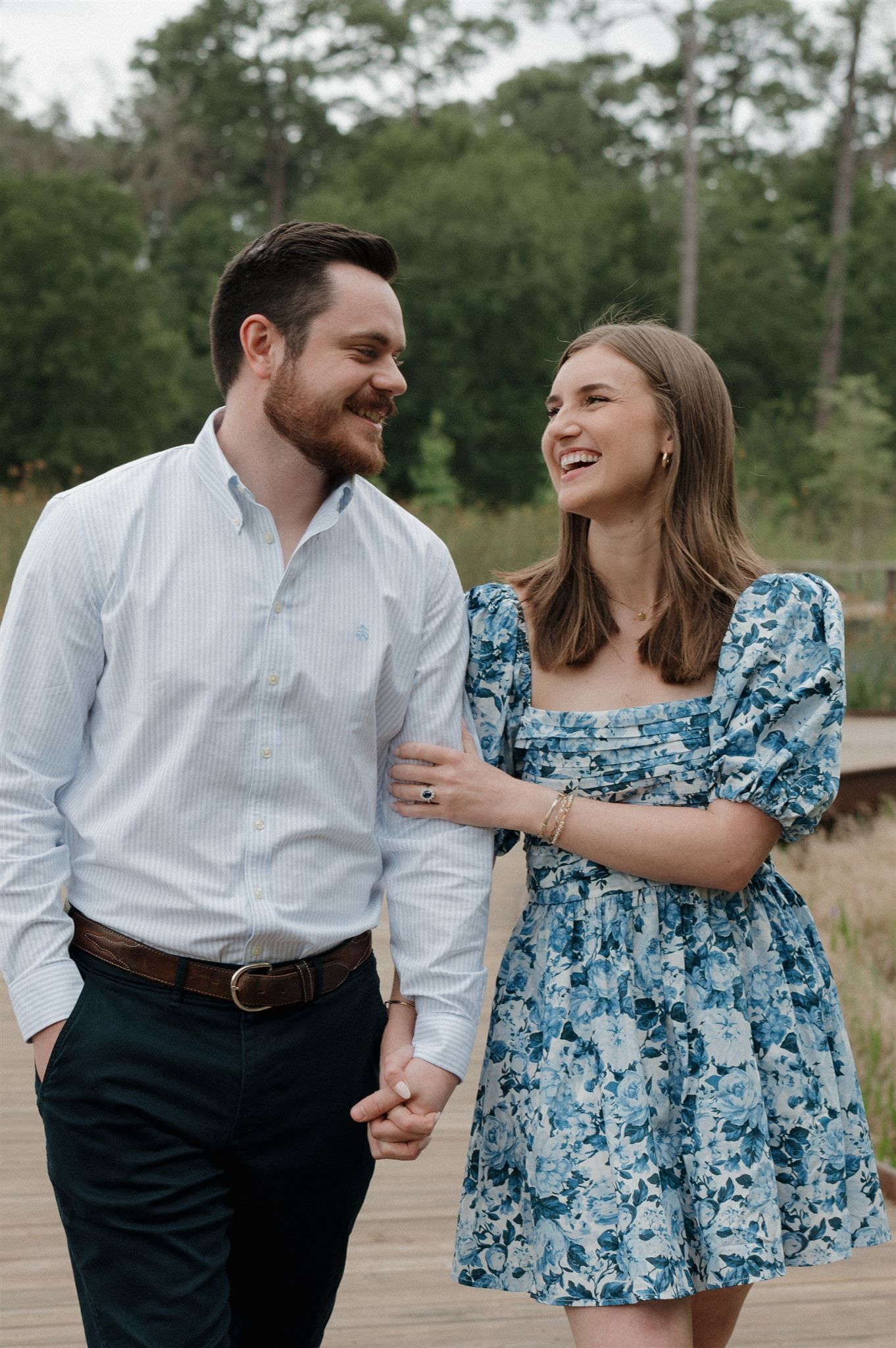 A couple in casual attire shares a joyful moment while walking on a wooden boardwalk surrounded by greenery.
