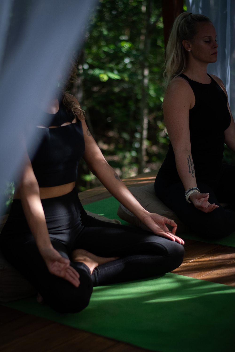People in black athletic wear meditating on green yoga mats in a sunlit space.