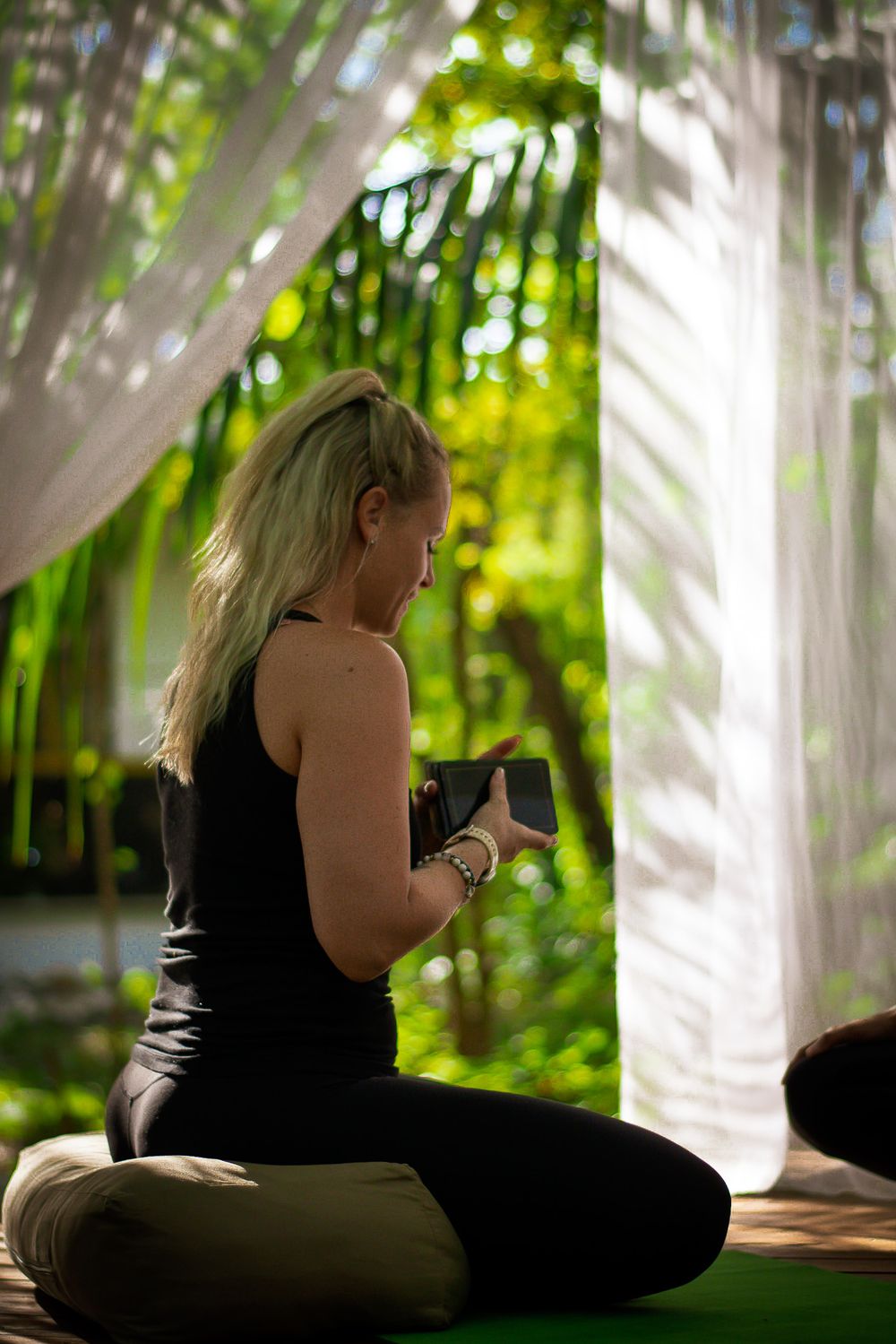 Person in dark activewear sitting cross-legged on yoga mat surrounded by white curtains.