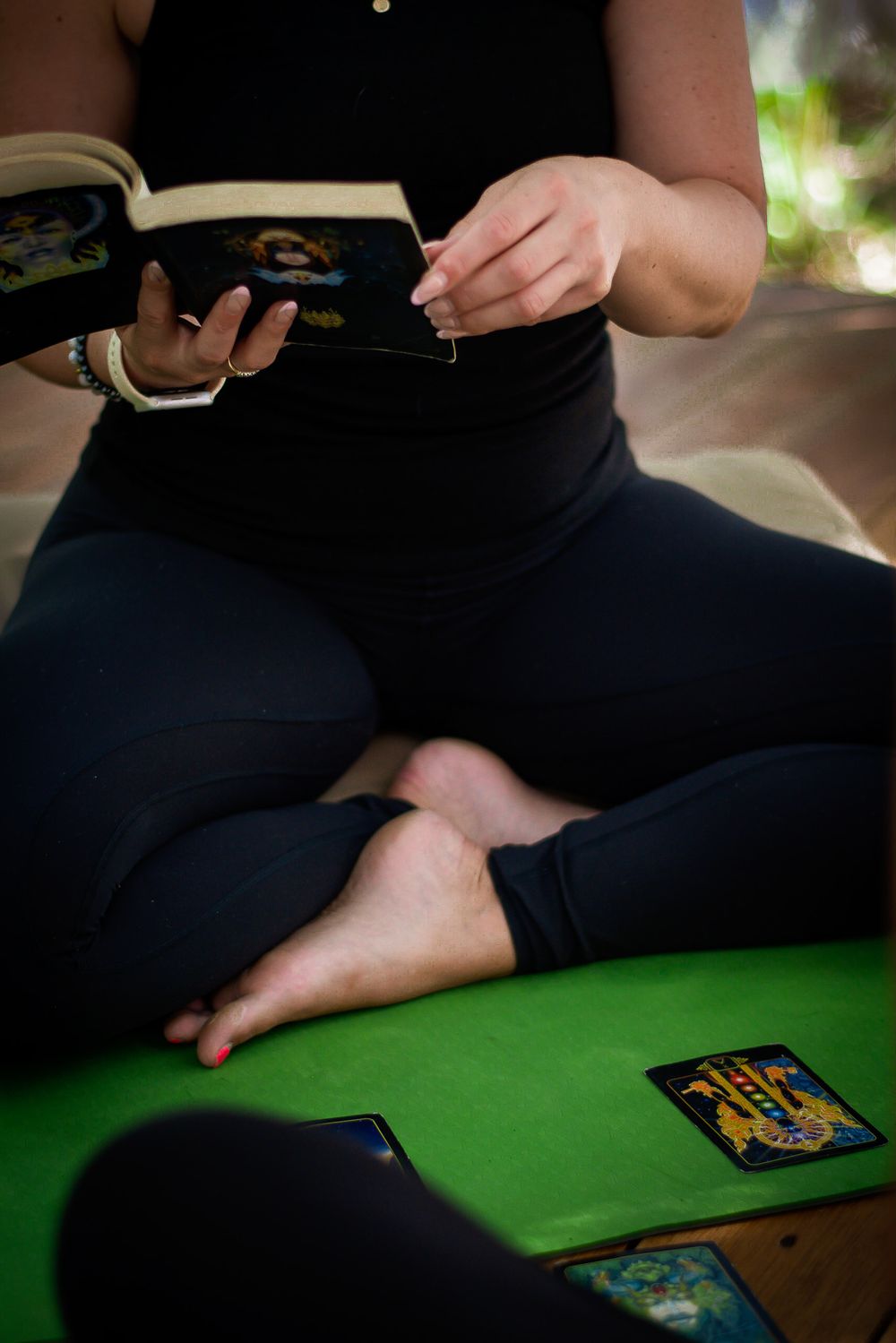 Hands holding a book while sitting cross-legged on a green surface with tarot cards.