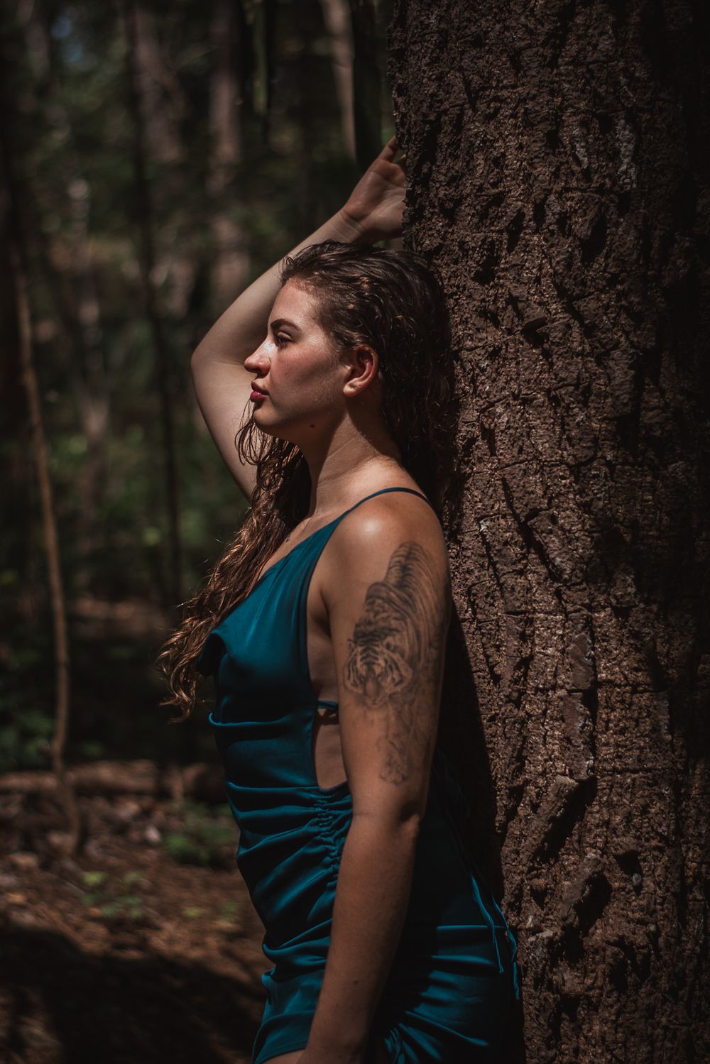 Person in teal dress leaning against tree trunk in moody forest setting.