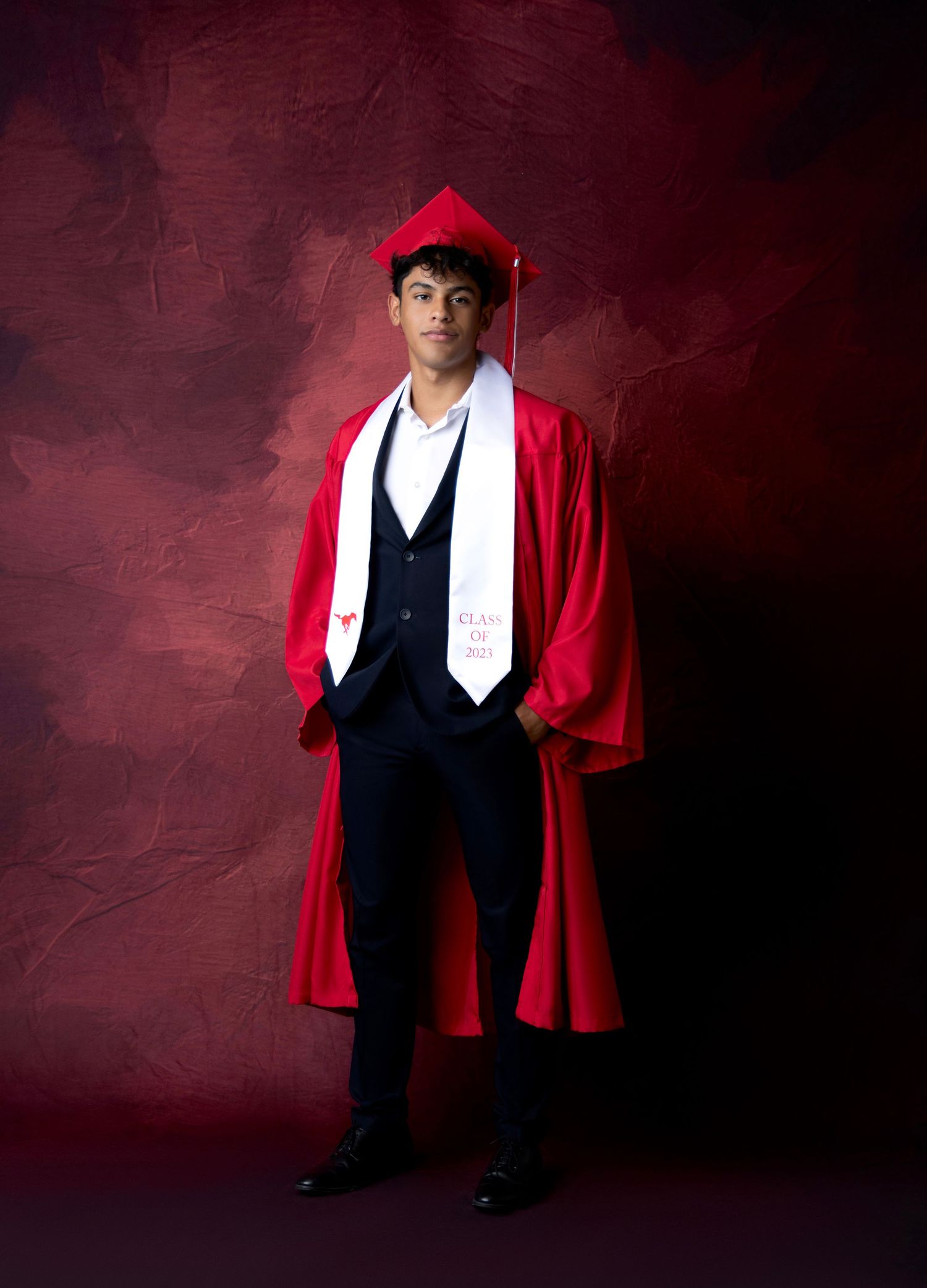 Graduate in black and red academic regalia poses against deep red background.