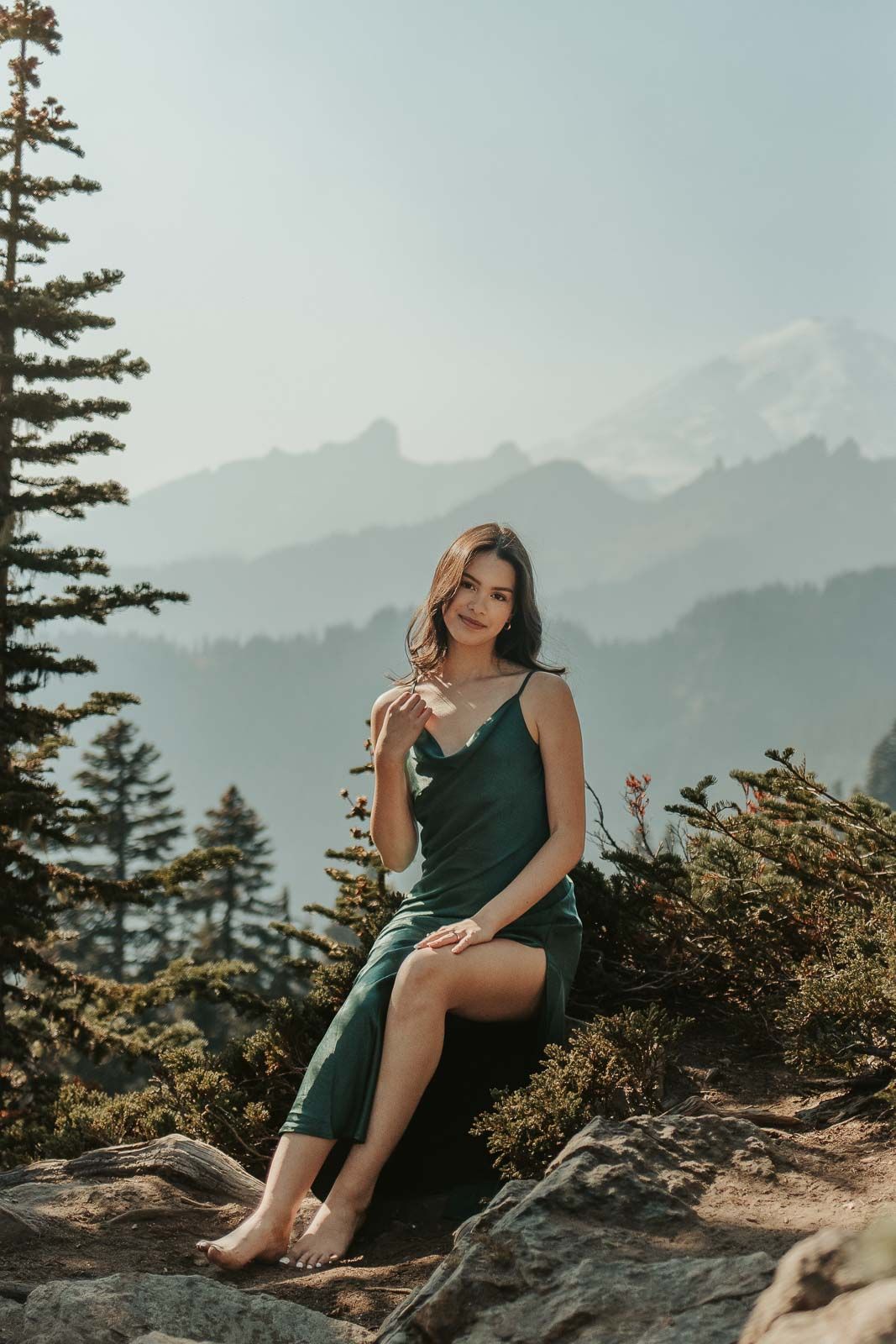 seated brown haired girl in long green silk dress in the mountains