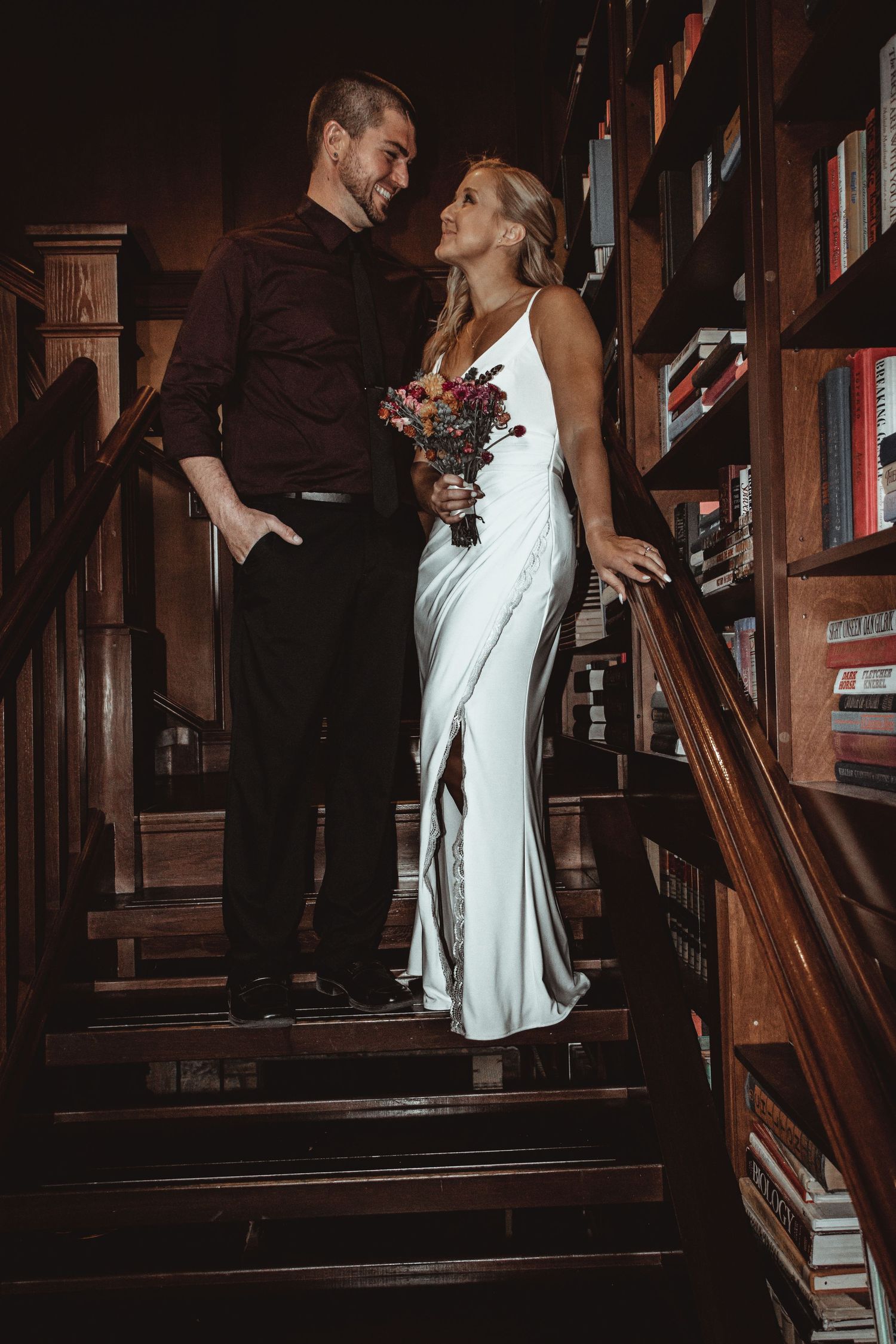 Romantic engagement photos on wooden library stairs with bookshelves in the background.