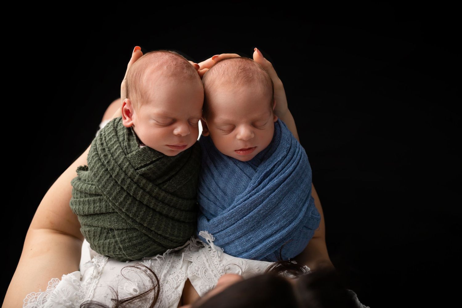 Two newborn babies wrapped in blue and green fabrics rest against each other on a dark background.