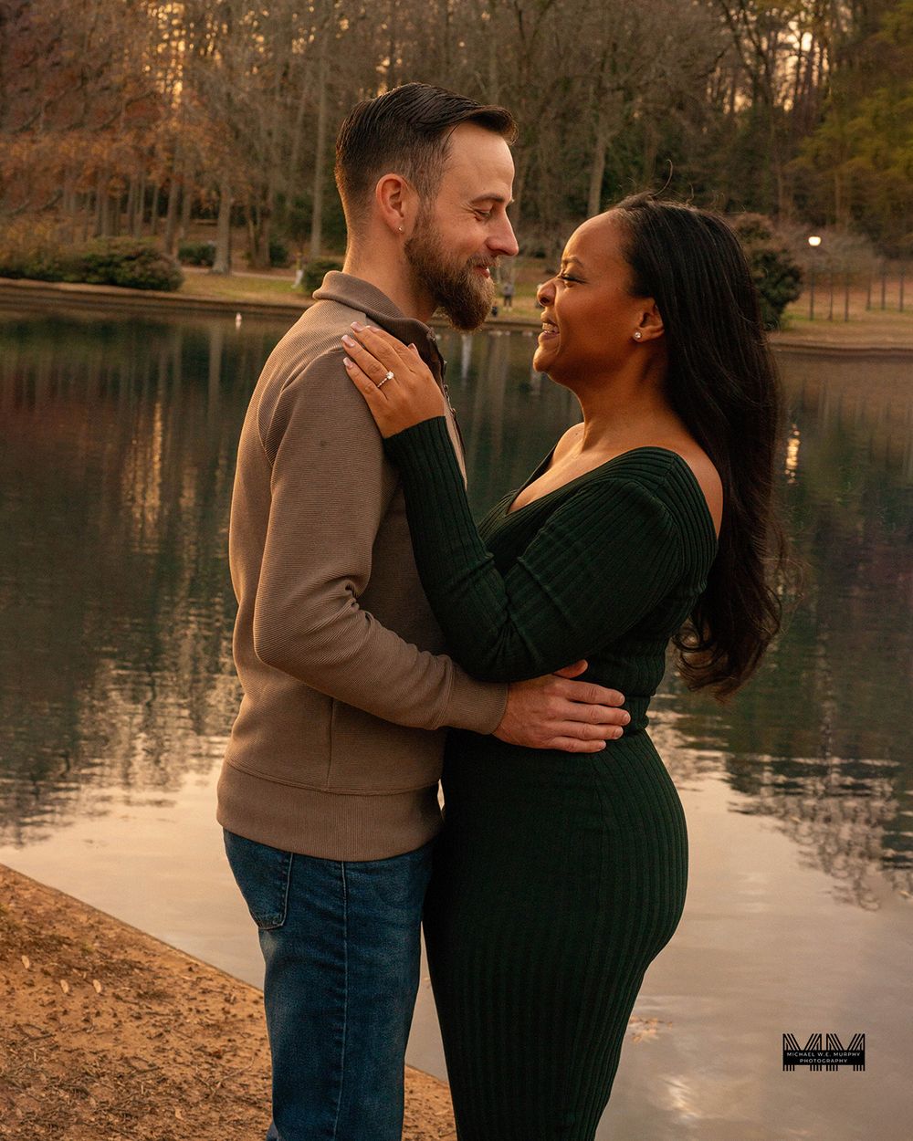 A romantic couple embraces by a serene lake during golden hour sunset in an autumn park setting.