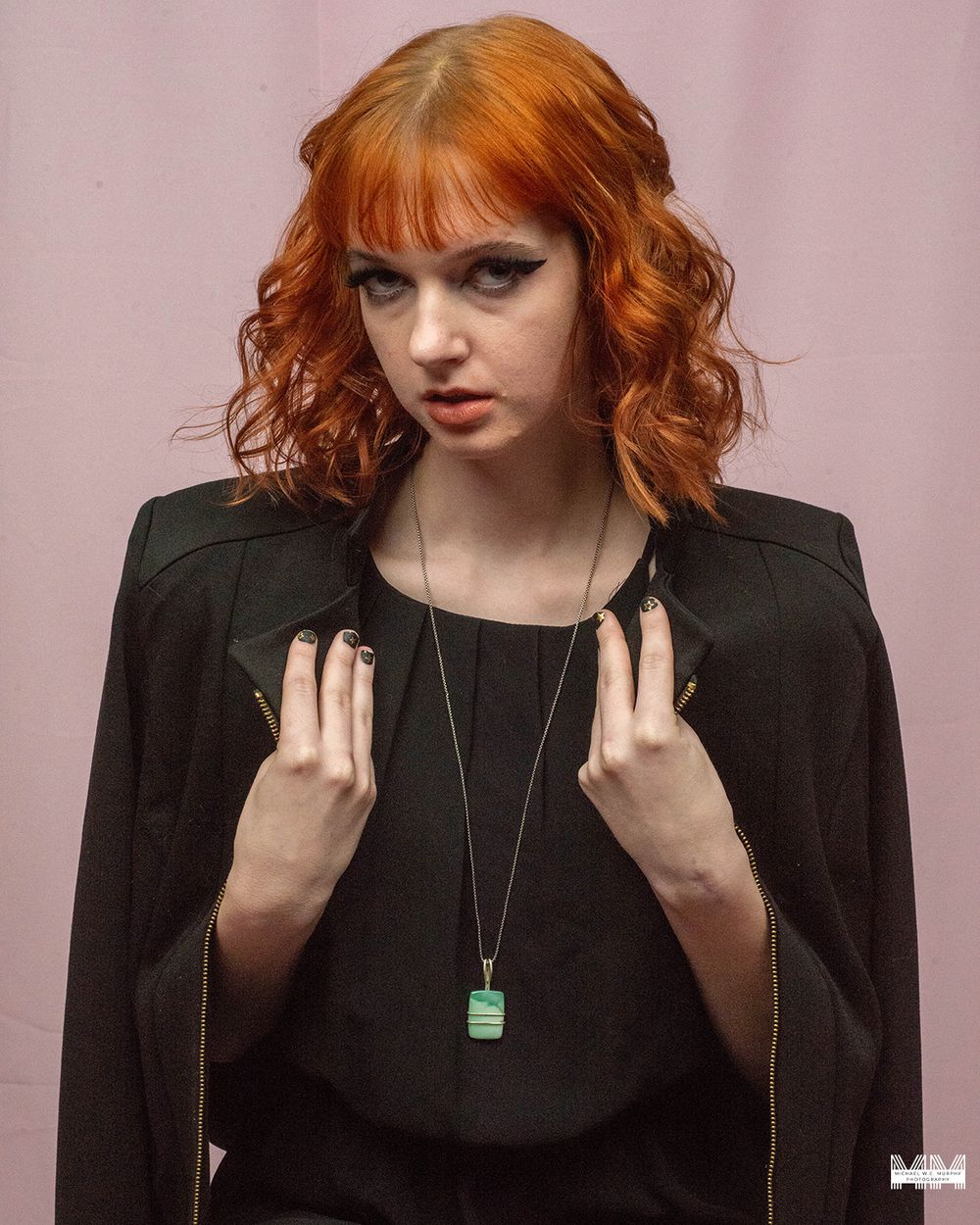 Woman with vibrant red curly hair wearing black jacket and green pendant necklace against pink background.