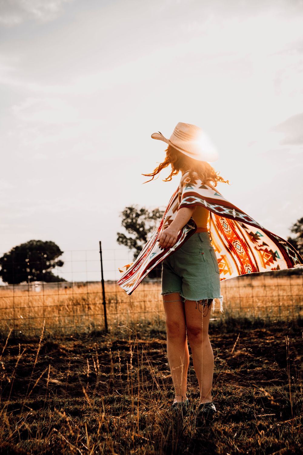 Silhouette spinning in sunlight wearing cowboy hat and flowing shawl in rustic field.
