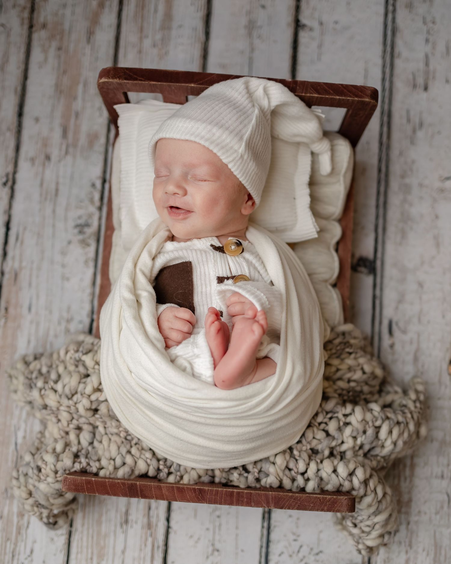 A newborn baby wearing white sleeps peacefully in a wooden basket on textured knit fabric.
