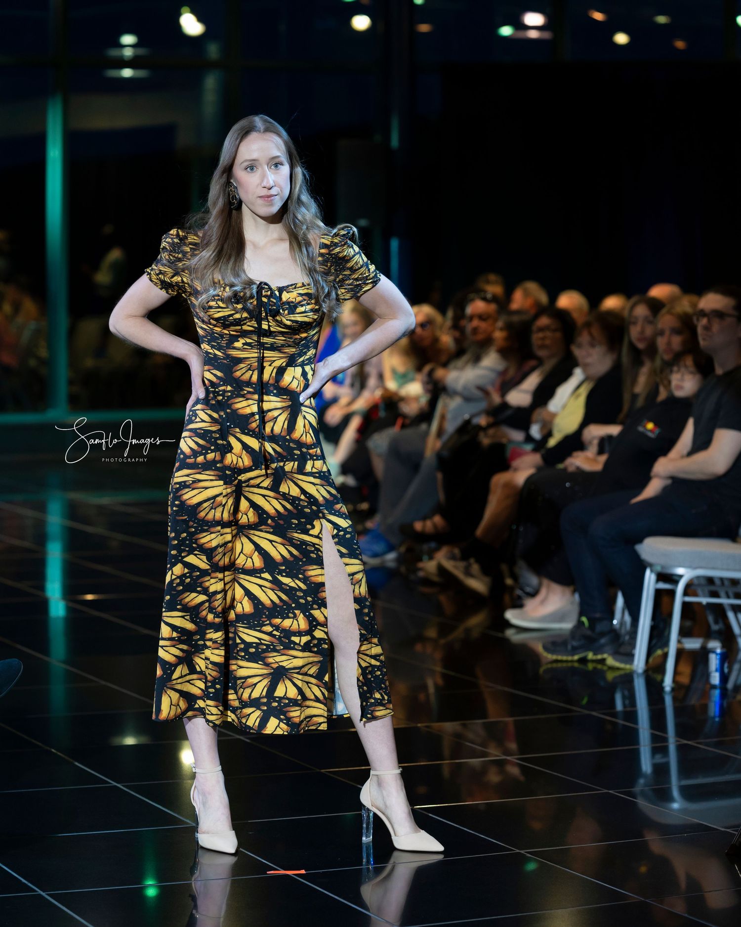 Model walks the runway in a black and gold butterfly print dress with white heels at a fashion show.
