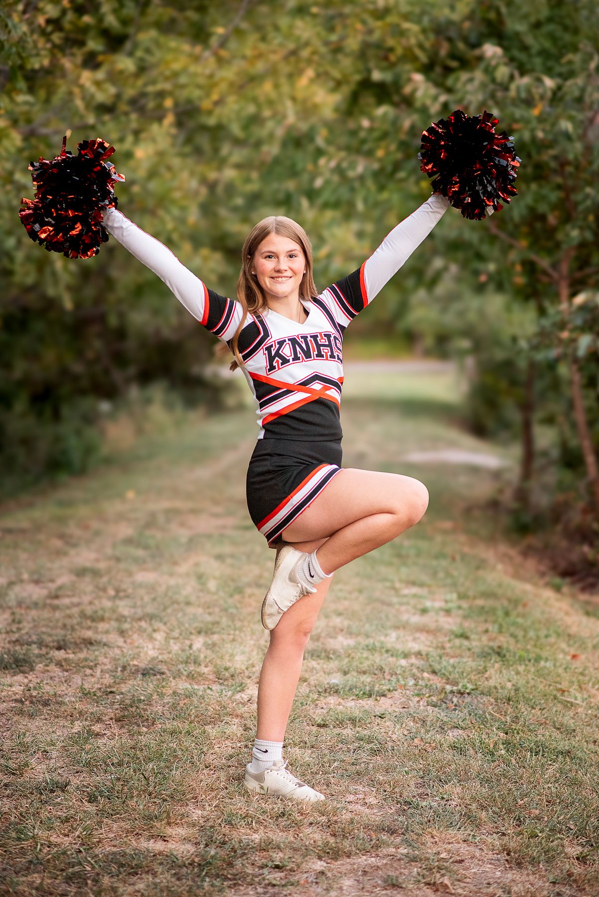High school senior photographer kansas city, cheerleader dressed in cheer uniform with pom poms posing in a cheer and smiling against trees