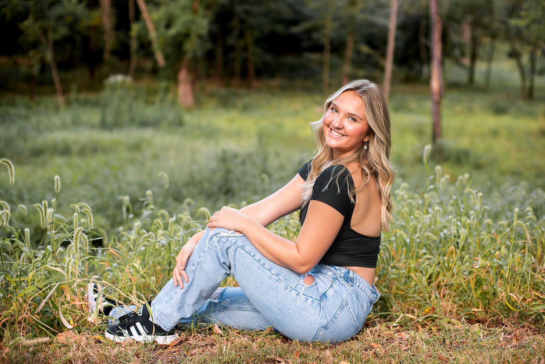 KC senior photographer, blond senior model posing in grassy area with flowers and golden sunlight wearing black tank top and jeans