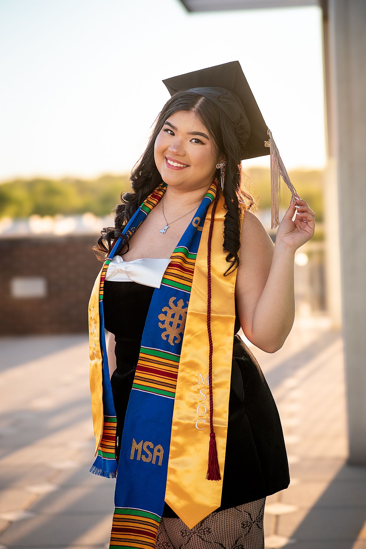 Smiling graduate in black cap, kente cloth stole, and MSA sash poses outdoors in golden hour light.