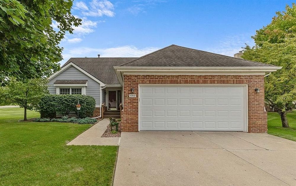 Brick and gray vinyl ranch home with two-car garage, concrete driveway, and mature trees.