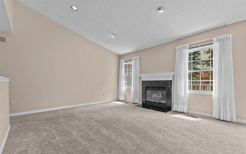 Bright living room with carpet, fireplace, white mantel, and large windows with white curtains.