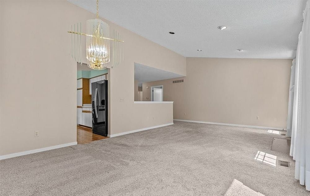Open carpeted living area with vaulted ceiling, chandelier, and view into kitchen with stainless appliances.