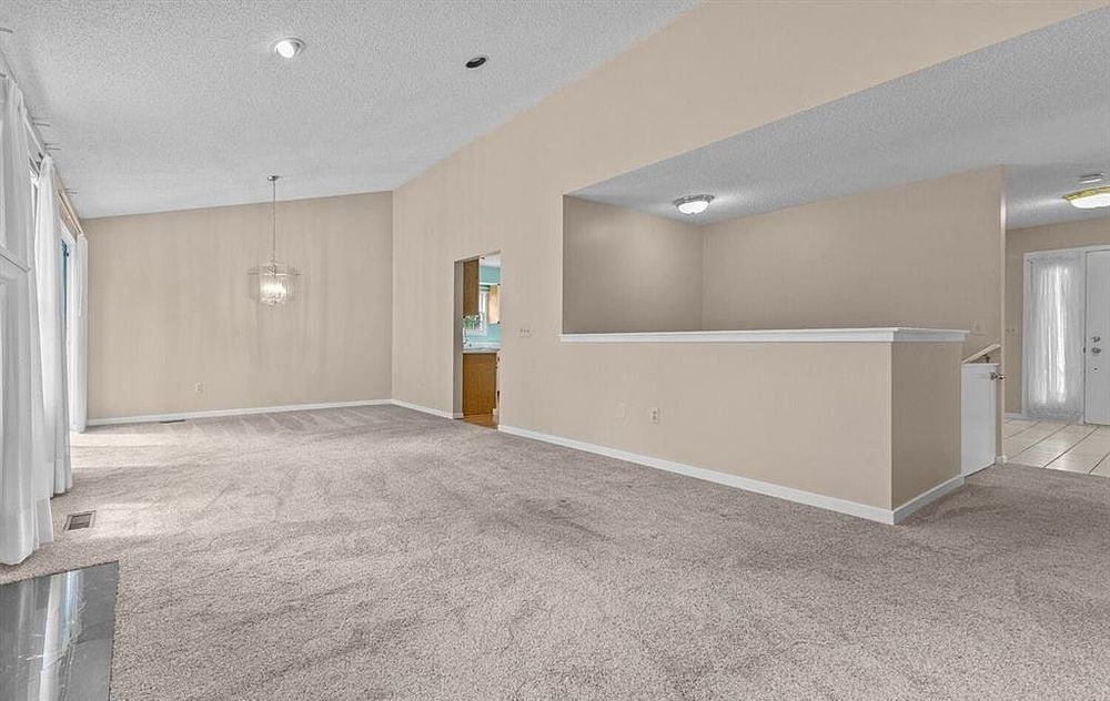 Open carpeted living space with vaulted ceiling, pendant light, half-wall divider, and neutral beige walls.