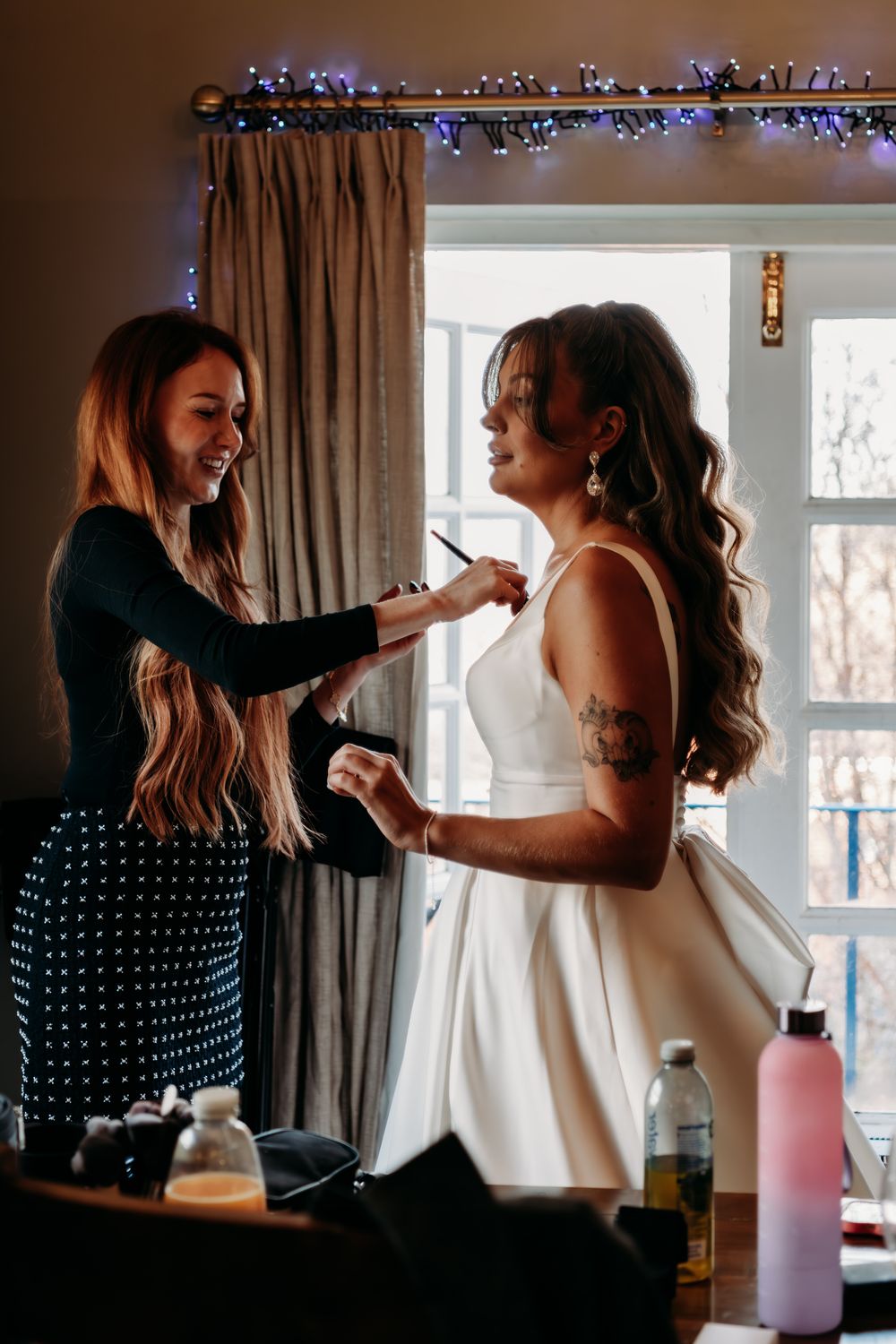 A makeup artist applies finishing touches to someone wearing a white wedding dress during bridal preparations.