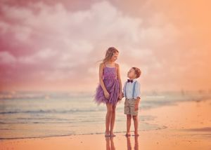 Two children in dressy outfits stand together on a pink-hued sunset beach.