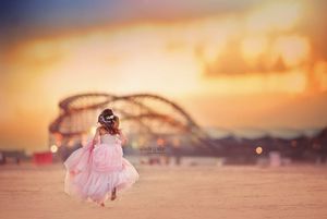 Little girl in pink tulle dress twirls near a roller coaster at sunset.