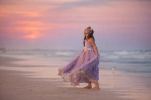 Young girl in lavender gown twirling on beach under vibrant pink and purple sunset sky.