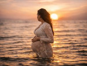 Pregnant woman in lace dress standing in ocean at golden sunset, cradling baby bump.