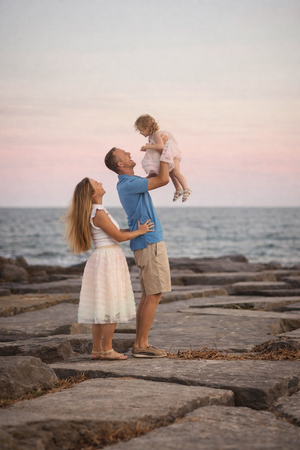 Happy family with father lifting toddler girl on rocky seaside at pink sunset.
