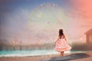 Little girl in pink dress gazes at colorful ferris wheel on dreamy beach.