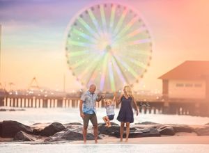 Family of three plays on rocky beach with glowing ferris wheel at sunset.