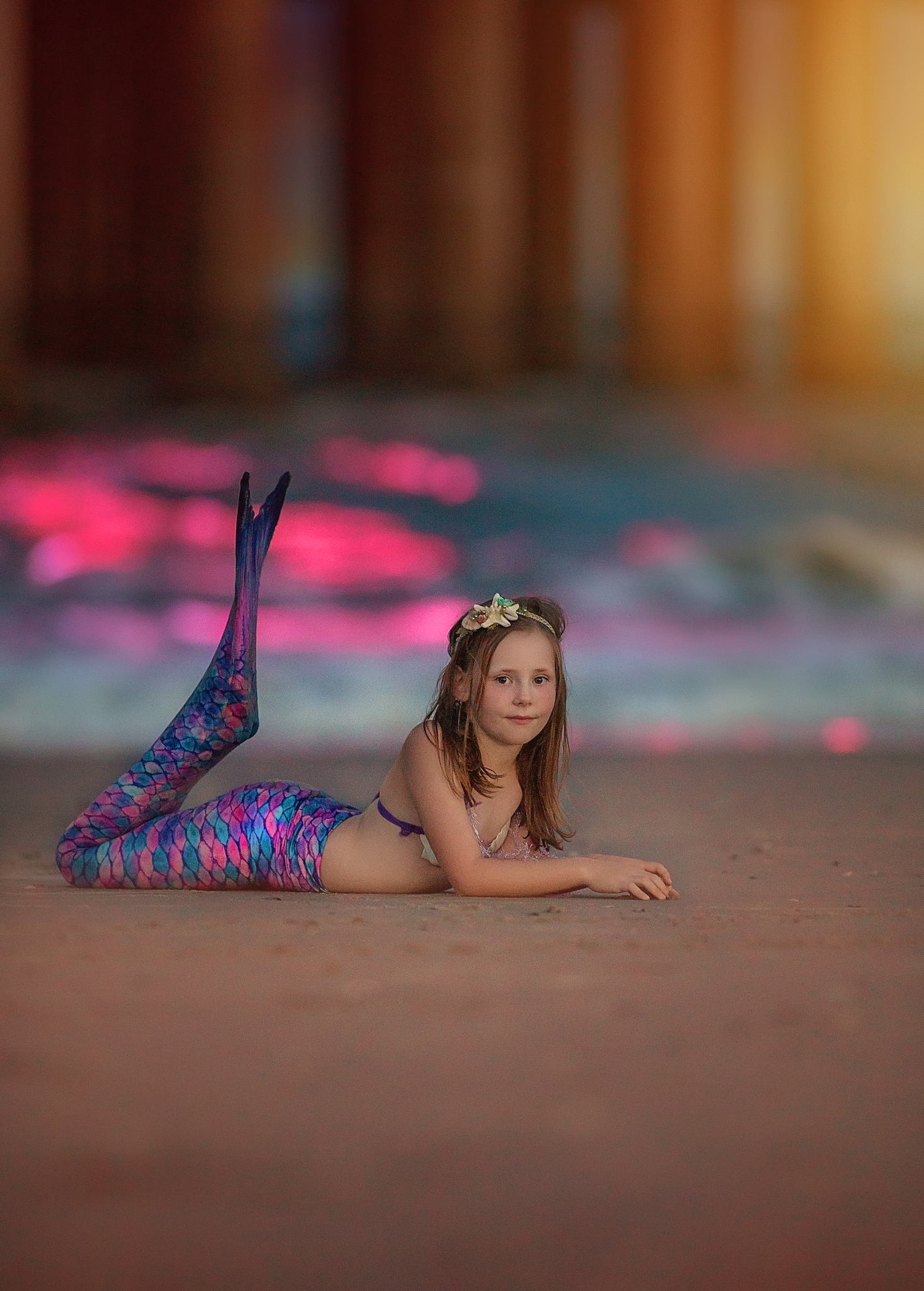 Young girl poses as a colorful mermaid on sandy beach with bokeh lights.