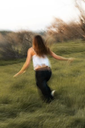 Young woman running through green grassy field, motion blur effect, wearing white top and jeans, trees in background.