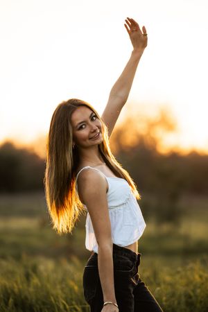 Young woman in white top and jeans posing at sunset in a field, raising her hand with golden backlight.