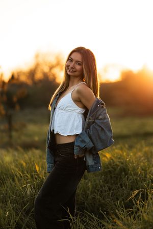 Young woman in white crop top and denim jacket posing in a field during golden hour sunset.