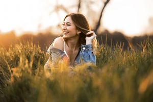 A smiling young woman sits in tall golden grass during sunset, wearing a denim jacket in a serene outdoor setting.