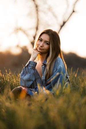 Young woman in denim jacket sitting in golden grass field at sunset, resting chin on hand, looking at camera.