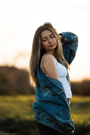 Young woman in denim jacket and white top posing in a golden sunset field with long flowing hair.