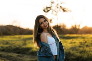 Young woman in denim jacket and white top poses in golden sunset field with long brown hair.
