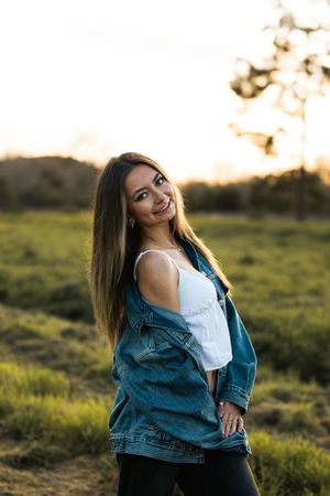 Young woman in denim jacket and white top smiling outdoors at golden hour sunset in green field.